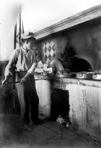 Φούρναρης, Κολωνάκι, μεταξύ 1915 και 1921, Φωτογραφικό Αρχείο Πέτρου Πουλίδη (ΕΡΤ): Το επάγγελμα του αρτοποιού ήταν ένα απ’ αυτά όπου το μοντέλο της μαζικής παραγωγής προϊόντων συνάντησε σοβαρές αντιστάσεις, ενώ τη δεκαετία του 1930 κατοχυρώθιηκε ως «κλειστό επάγγελμα». Στην αντοχή των βιοτεχνών έπαιξαν ρόλο διάφοροι παράγοντες. Η διατίμηση του ψωμιού κρατούσε αρχικά τα κέρδη στον κλάδο σε χαμηλά επίπεδα, αποθαρρύνοντας τις μεγάλες επενδύσεις. Οι καταναλωτές προτιμούσαν την ποιότητα και την ποικιλία του ψωμιού που παραγόταν βιοτεχνικά. Το ψωμί πουλιόταν συνήθως με πίστωση, και ο βερεσές ήταν ένα σύστημα που βασιζόταν στην προσωπική γνωριμία και μπορούσε να λειτουργήσει μόνο στη μικρή κλίμακα της γειτονιάς. Οι αρτοποιοί για να αντεπεξέλθουν στον ανταγωνισμό δούλευαν περισσότερες ώρες και αποδέχονταν ένα χαμηλότερο βιοτικό επίπεδο, κατοικώντας για παράδειγμα στον φούρνο ώστε να μην πληρώνουν νοίκι. Τέλος μπορούσαν να υπολογίζουν σε κάποια έξτρα έσοδα από «τα ψηστικά», την αμοιβή τους όταν έψηναν στον φούρνο τους το ψωμί ή το φαγητό που τους πήγαιναν οι νοικοκυρές, όπως φαίνεται ότι συμβαίνει στη φωτογραφία. © Πανεπιστημιακές Εκδόσεις Κρήτης.